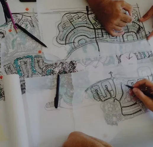 Several architectural sketches and plans are spread across a table, with hands holding pens and making annotations on the designs.