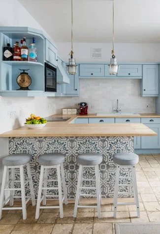 Bright kitchen featuring light blue cabinetry, a wooden countertop, bar stools, and decorative tile, with drinks displayed on shelves.