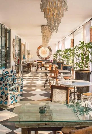 Bright, stylish café with checkered floors, eclectic furniture, greenery, and unique light fixtures, creating a welcoming atmosphere.