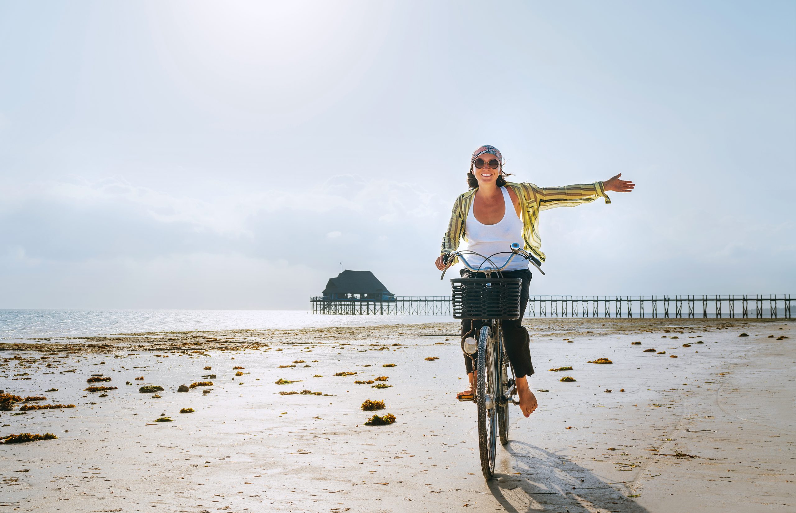 • Woman cycling barefoot on a Zanzibar beach at low tide, smiling with arms outstretched and traditional jetty in the background • Carefree woman riding bike on tropical beach in Zanzibar, showcasing relaxed coastal lifestyle and remote living potential • Young woman enjoying slow living in Zanzibar, cycling on beach with overwater bungalow in background
